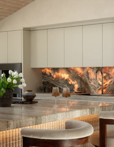 Modern kitchen with beige cabinets, illuminated stone backsplash, marble countertop, two chairs, and decorative items including a vase of white tulips.