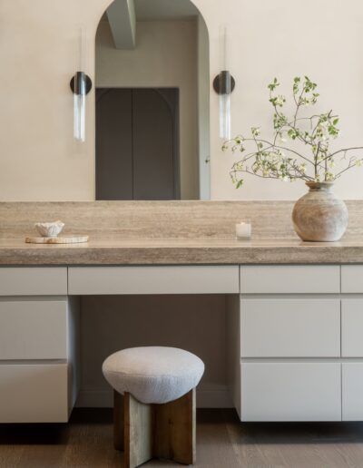 Minimalist vanity with a stone countertop, large arched mirror, modern sconces, a ceramic vase with branches, a small candle, and a cushioned wooden stool.