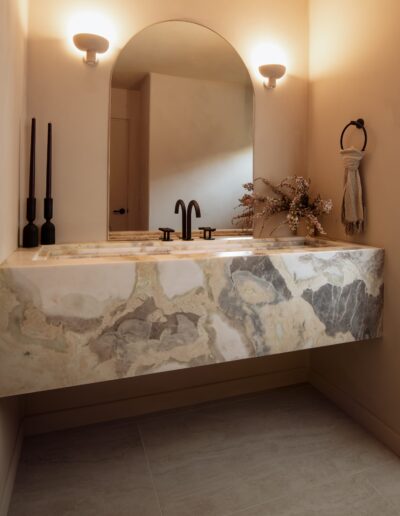 Modern bathroom with a marble-patterned floating vanity, black faucet, arched mirror, two wall sconces, candles, and a small floral arrangement on the right.